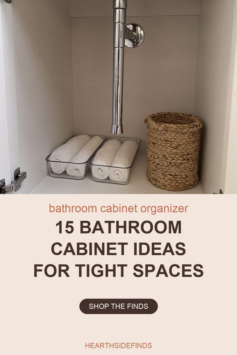 Photo looking into the open cabinet under a bathroom sink, with white melamine walls and floor and a chrome drain pipe running down the centre from the top of the frame past a round chrome wall fitting behind it. Two low clear plastic bins sit side by side on the cabinet floor, each holding four white washcloths rolled and laid on their sides so the spiral ends face the camera. A tall round basket woven from pale straw stands to their right. Silver cabinet hinges show at both side edges of the frame.