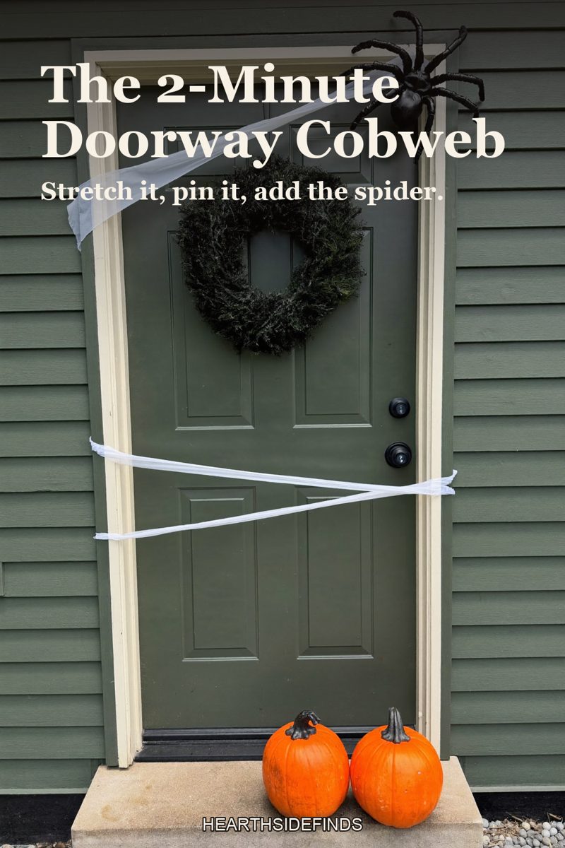 Green front door with a plain wreath, white stretchy spider webbing across it, a large black spider at the top corner and two orange pumpkins on the step.