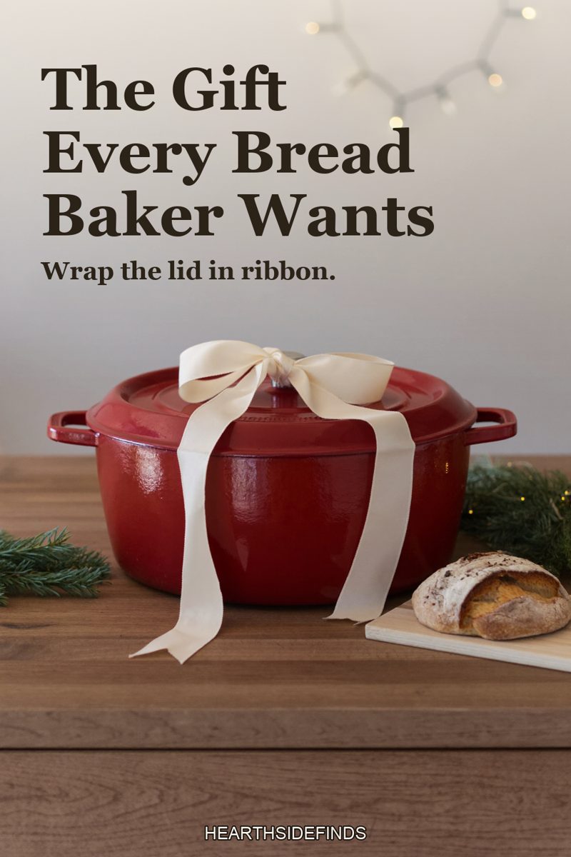 Red enameled dutch oven tied with a cream ribbon beside a sourdough loaf on a kitchen counter.