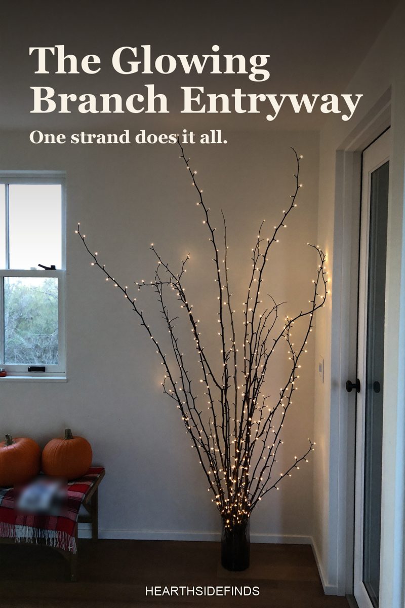 Floor vase of bare branches wrapped in warm fairy lights beside an entry bench with pumpkins.