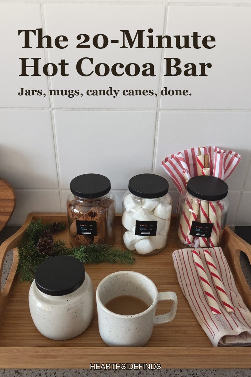 Wooden tray on a kitchen counter with glass jars of marshmallows and cocoa with black lids, speckled stoneware mugs, candy canes and pine sprigs, white tile behind.