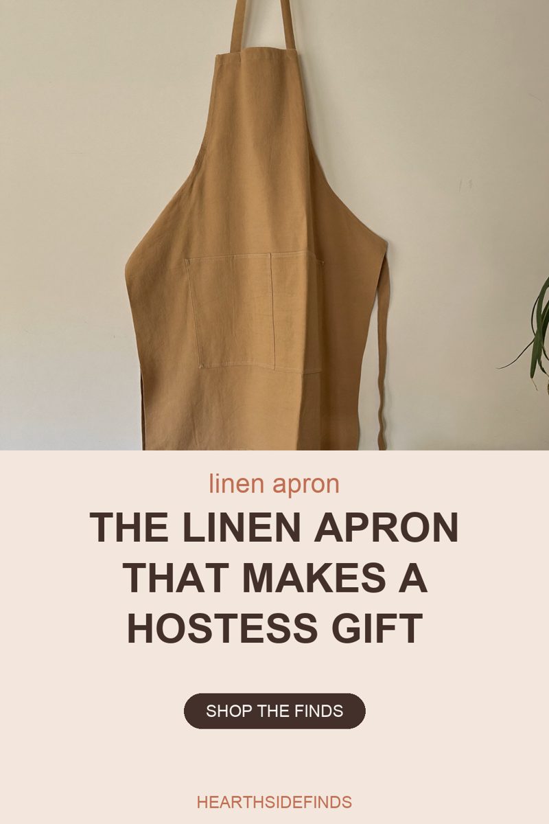 Photo of a camel-coloured linen apron hanging flat against a plain off-white wall, its neck strap running up out of the top of the frame and one waist tie falling loose down the right-hand side. A large square patch pocket is stitched across the front at waist height, the linen falls in soft creases, and a few green leaves of a houseplant reach in at the right edge.