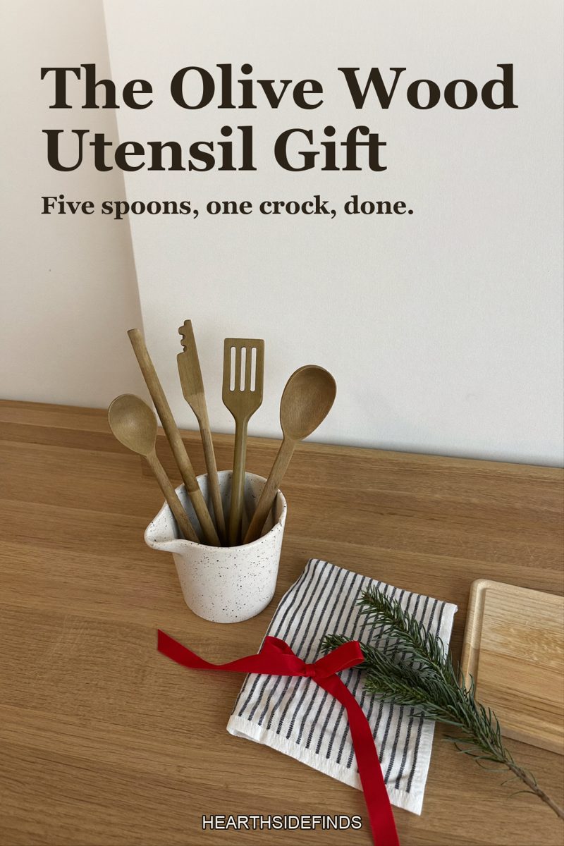 Olive wood spoons and spatulas standing in a white speckled stoneware crock on a light oak counter, a striped tea towel with a red ribbon and a wooden board beside it.