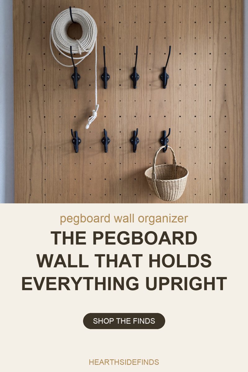 Photo of a wood pegboard panel filling the frame, its grid of small drilled holes running edge to edge, mounted on a pale grey wall. Eight black metal double hooks are screwed into the board in two rows of four. A coil of cream cotton rope hangs from the top left hook with one loose end trailing down the board into a small knot, and a shallow basket woven from pale straw hangs by its handle from the right-hand hook of the lower row. The other six hooks are empty.