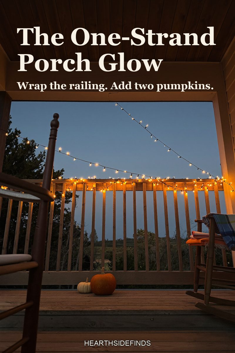 Wooden front porch at dusk with a strand of warm fairy lights along the railing, pumpkins on the step and a plaid blanket on a rocking chair.
