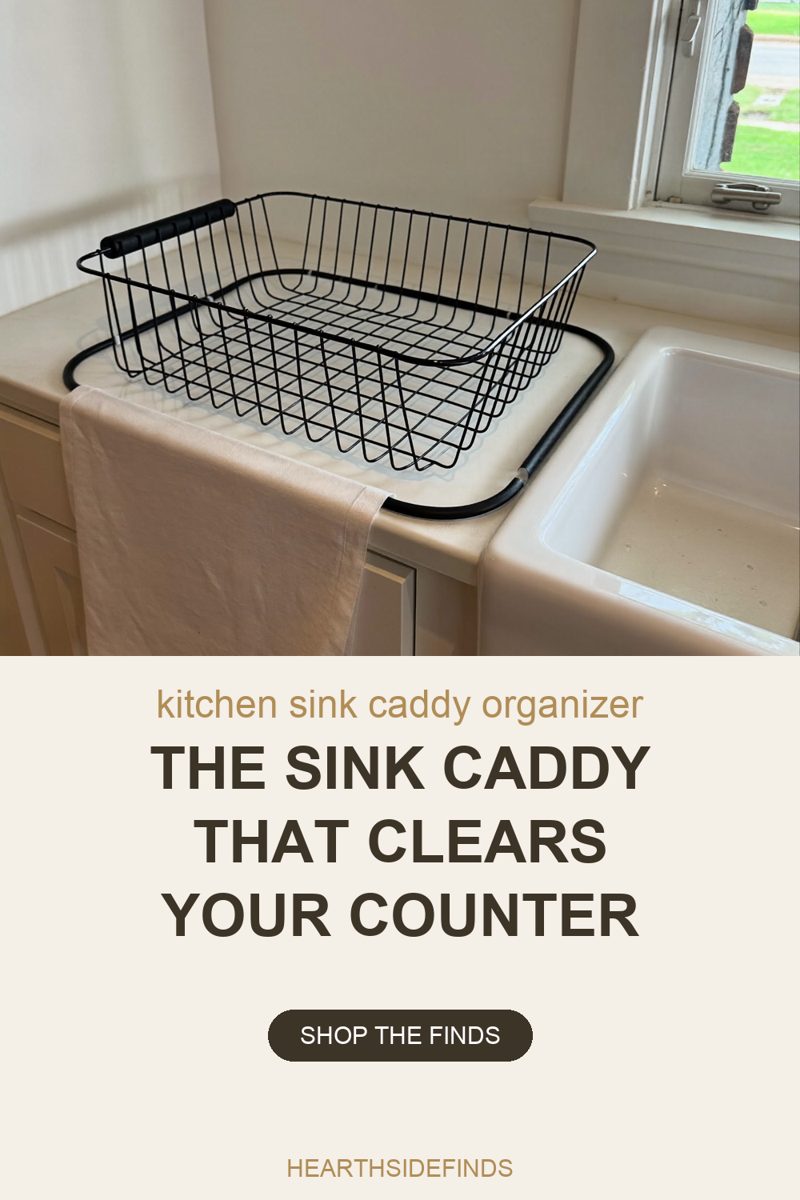 Photo of a bright kitchen corner lit by a window at the upper right. An empty rectangular basket of thin black wire, with a black moulded grip handle across its near long side, sits on a pale countertop inside a slim black rounded-rectangle tube frame that runs around it and down to the rim of a white apron-front sink at the right. A cream linen towel hangs over the counter edge and down the plain pale cabinet doors below.