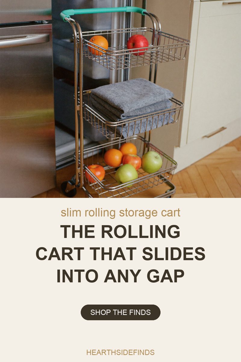 Photo of a narrow three-tier chrome wire rolling cart with a mint-green grip handle standing in the gap between a stainless steel refrigerator and a run of cream cabinets, its top basket holding an orange and a red apple, its middle basket a stack of folded grey towels and its bottom basket more oranges and green apples, with one black castor visible on herringbone wood floor