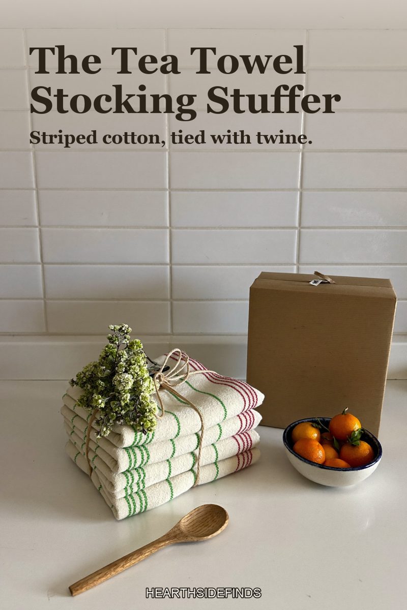 Stack of folded cream cotton tea towels with red and green stripes tied with twine on a white counter, a sprig of holly, a wooden spoon, a bowl of clementines and a kraft box beside them.