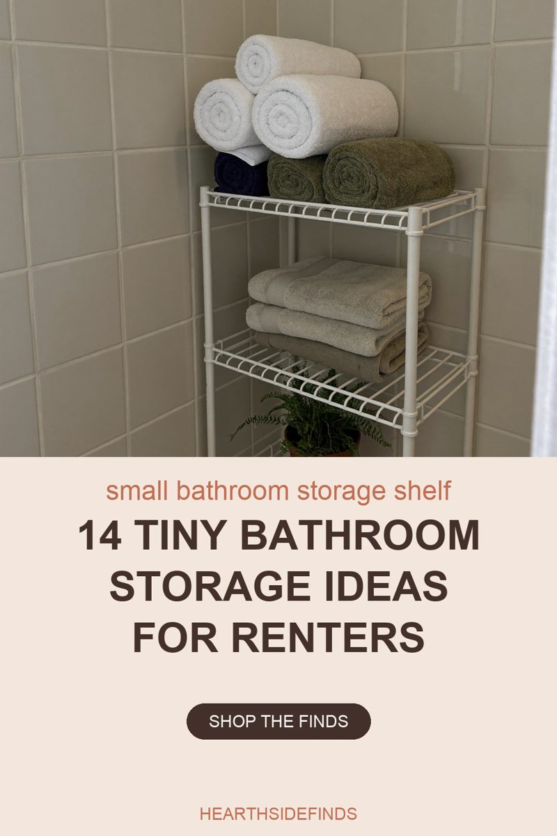 Photo of a white wire freestanding shelf unit standing in the corner of a bathroom tiled in matte greige square tile. Six rolled towels on the top tier: one navy and two olive green resting on the wire, with three white ones stacked on top of them. Three folded pale sage towels stacked on the tier below. The green fronds of a potted fern show through the wire of a lower shelf at the bottom edge of the frame. A pale curtain edge runs down the right side.