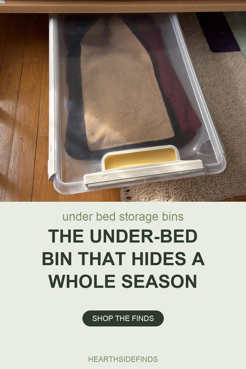 Photo looking down at a long clear plastic under-bed storage box, lid closed, pulled out from under a light wood bed frame onto a wood plank floor. Three folded items show through the lid: a charcoal one filling the left side, a camel fleece folded on top of it down the middle, and a dark maroon one along the right. A pale grey latch flap with a yellow inner panel sits at the front edge of the lid. A cream shag rug fills the right side, with a navy and green striped rug edge at the top right corner.